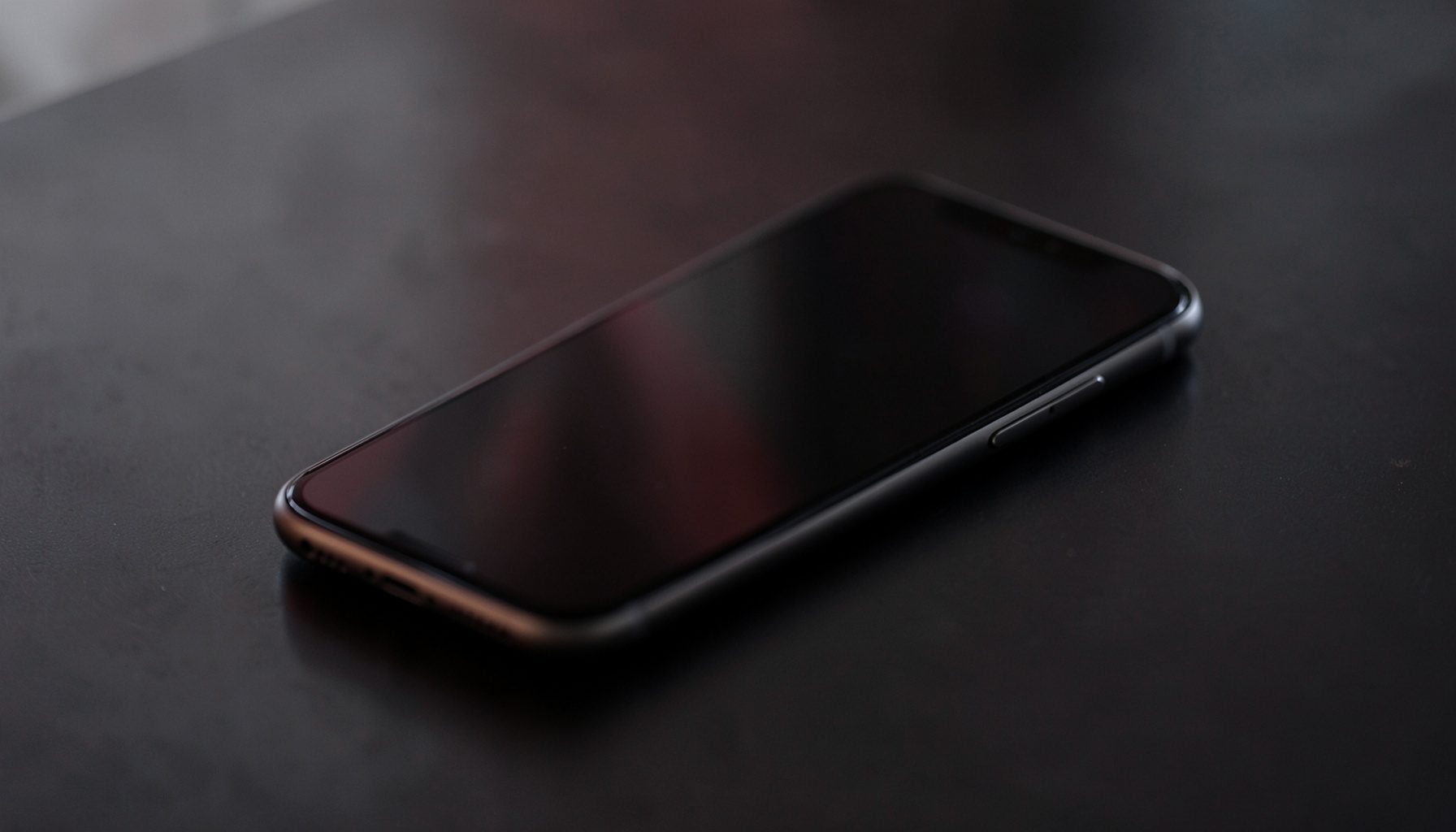 A smartphone resting on a dark desk surface with soft reflections, moody charcoal and deep red tones