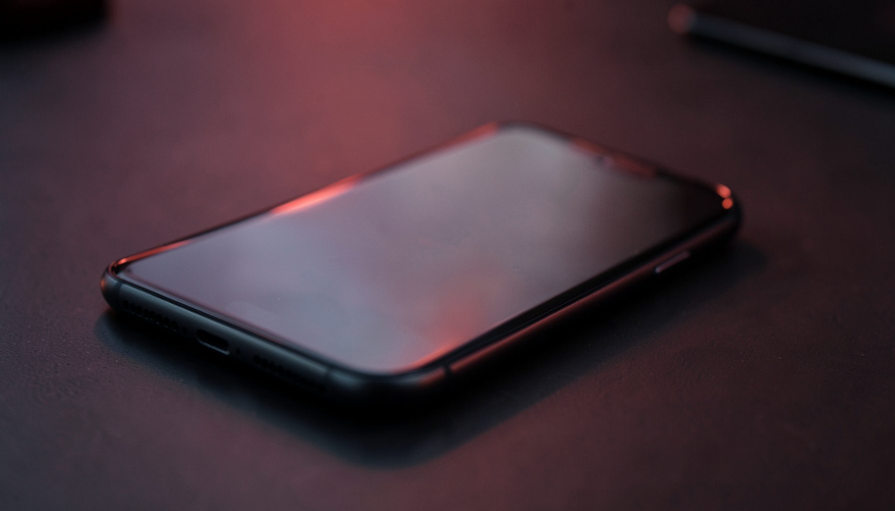 Close-up of a modern smartphone resting on a dark desk surface with soft red and charcoal ambient lighting